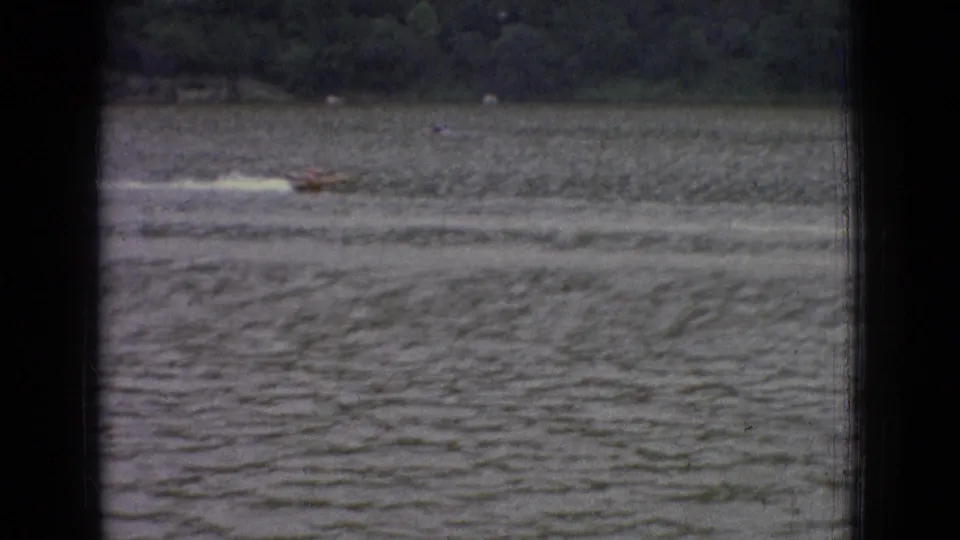 1965 New Jersey 8mm frame with a small motorboat traveling across rippled water, its pale wake stretching behind it toward the wooded shore.