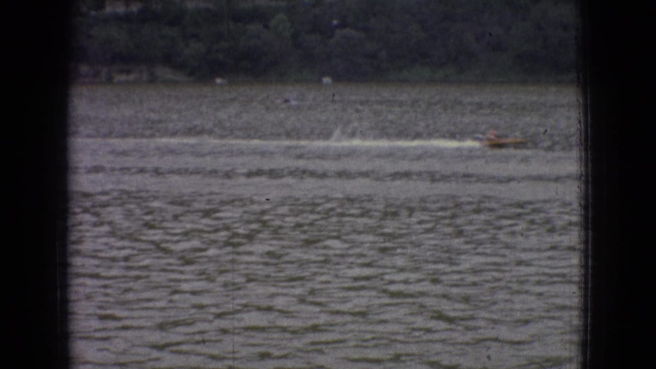 1965 New Jersey 8mm frame showing a small speedboat moving across choppy water with a wooded shoreline in the distance and dark vertical framing at the edges.