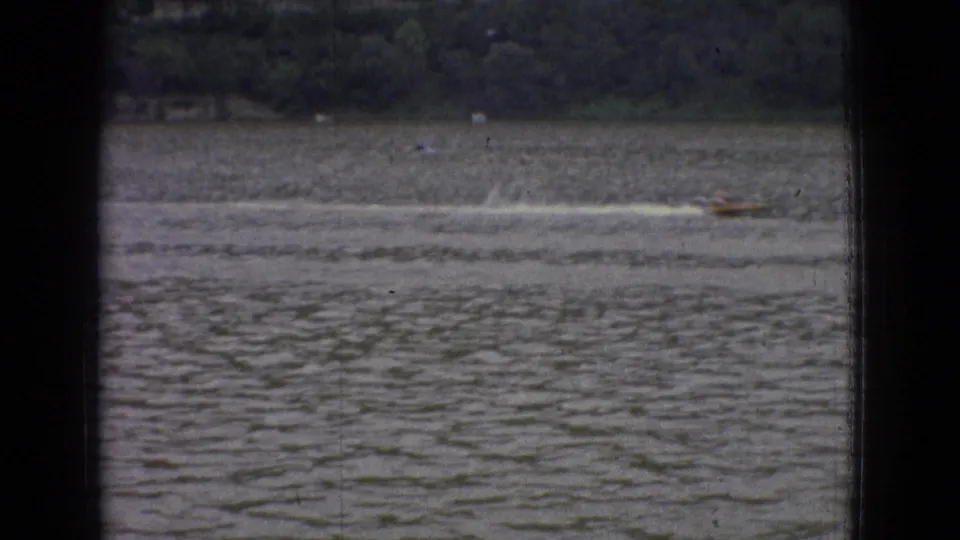 1965 New Jersey 8mm frame showing a small speedboat moving across choppy water with a wooded shoreline in the distance and dark vertical framing at the edges.