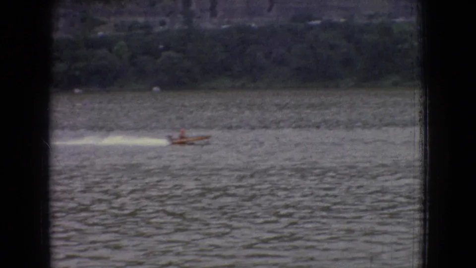 1965 New Jersey home-movie frame with a small motorboat traveling at speed across rippled water, leaving a bright wake below a dark wooded shoreline.