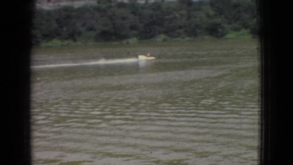 1965 New Jersey 8mm frame showing a small speedboat near the lower center of the view, trailing a long white wake across a broad river or lake with trees behind it.