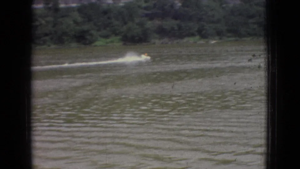 1965 New Jersey home-movie frame with a small boat partly softened by motion blur, a wide white wake, choppy foreground water, and a dark wooded shoreline beyond.