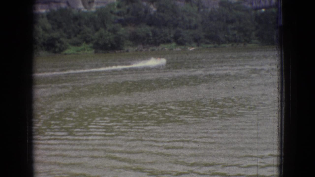 Small speedboat crossing a river near a wooded bank and rocky slope.