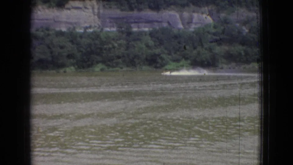 Distant motorboat in 1965 New Jersey home-movie footage, skimming across the river with a bright wake below trees and exposed rock.