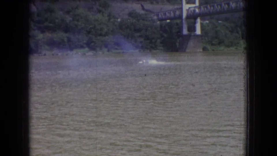 8mm frame of river racing action in New Jersey, with a speedboat wake in the midground and a bridge structure visible near the wooded shore.
