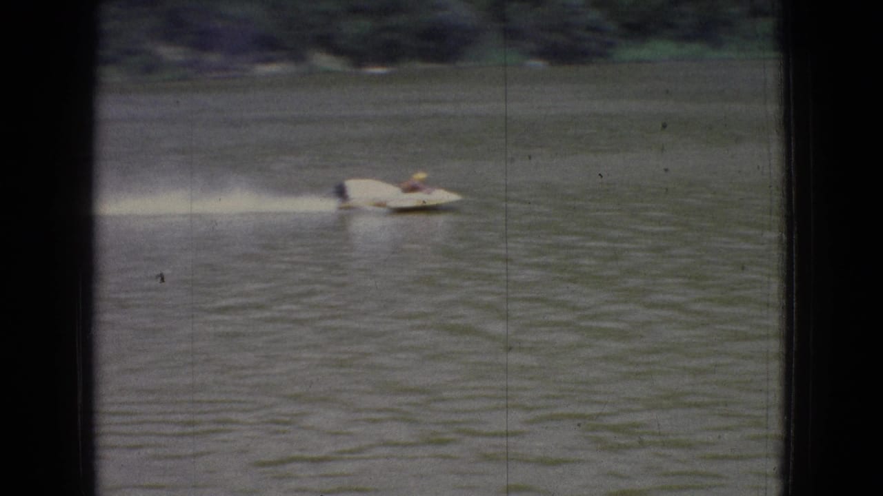 A white speedboat crosses a rippled New Jersey lake in 1965 home-movie footage, seen through dark vertical and horizontal framing bars with shoreline in the distance.