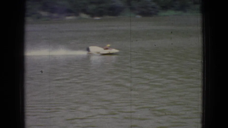 A white speedboat crosses a rippled New Jersey lake in 1965 home-movie footage, seen through dark vertical and horizontal framing bars with shoreline in the distance.