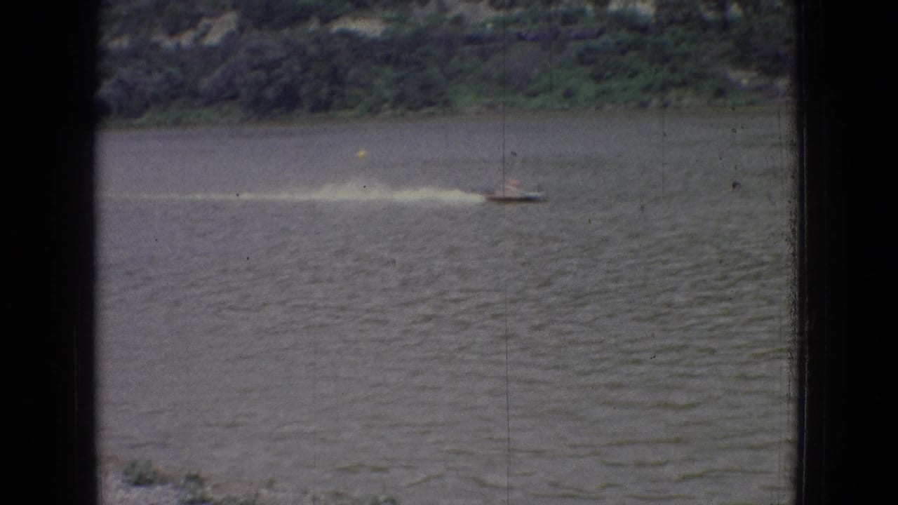 A distant small speedboat leaves a pale wake across the lake near a tree-lined shore in the 1965 New Jersey home-movie scene.
