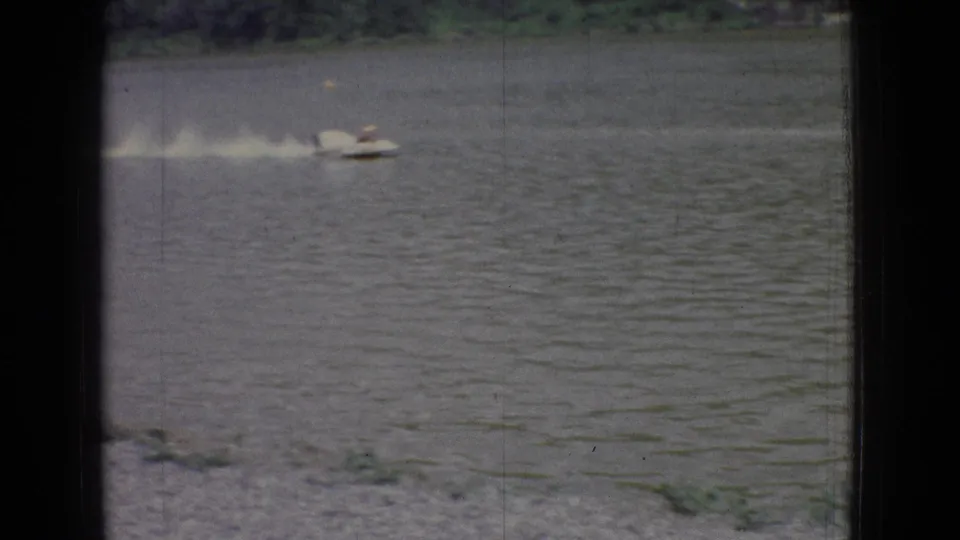 A white speedboat moves across the lake with a long pale wake, partially framed by dark bars and soft 8mm grain in the 1965 New Jersey footage.