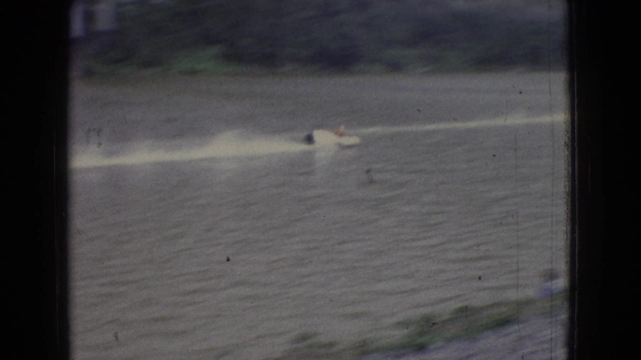1965 New Jersey 8mm frame showing a small motorboat crossing a broad river and leaving a bright wake near a wooded bank.