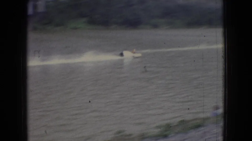 1965 New Jersey 8mm frame showing a small motorboat crossing a broad river and leaving a bright wake near a wooded bank.