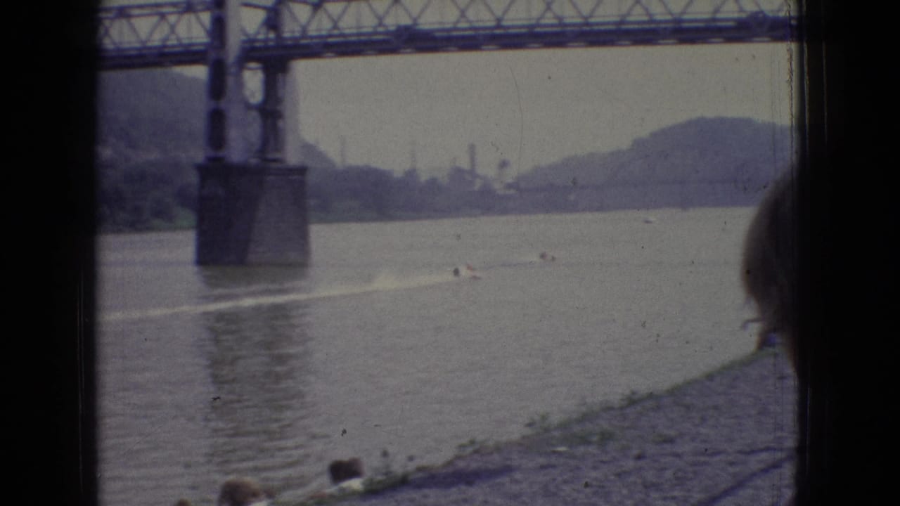 1965 New Jersey 8mm frame showing a steel truss bridge over the river, a distant boat wake, wooded hills, and a partial person visible at the right edge.