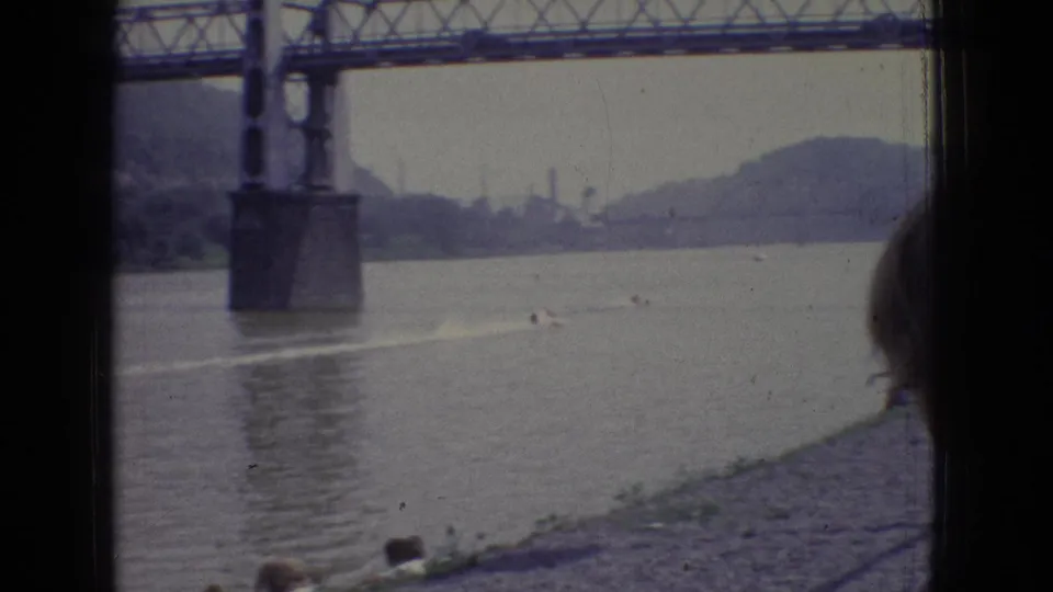 1965 New Jersey 8mm frame showing a steel truss bridge over the river, a distant boat wake, wooded hills, and a partial person visible at the right edge.