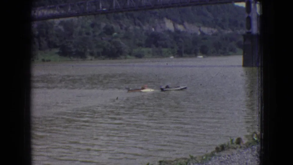 1965 New Jersey 8mm frame showing small boats on the river below a steel bridge, with wooded shoreline and bridge supports in the background.