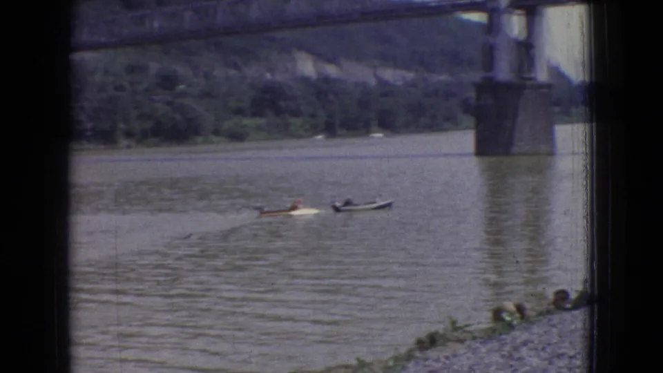 1965 New Jersey 8mm frame showing two small boats moving across the river beneath a steel bridge, with rocks and low vegetation along the near shore.