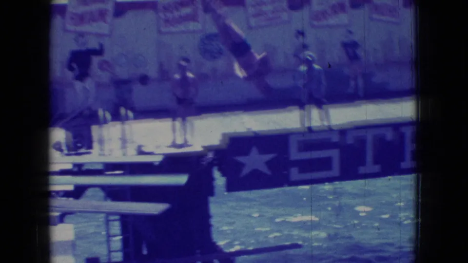 1965 New Jersey waterfront home-movie frame showing a partial sign with a star and the letters STE, water behind it, and several small human figures nearby.