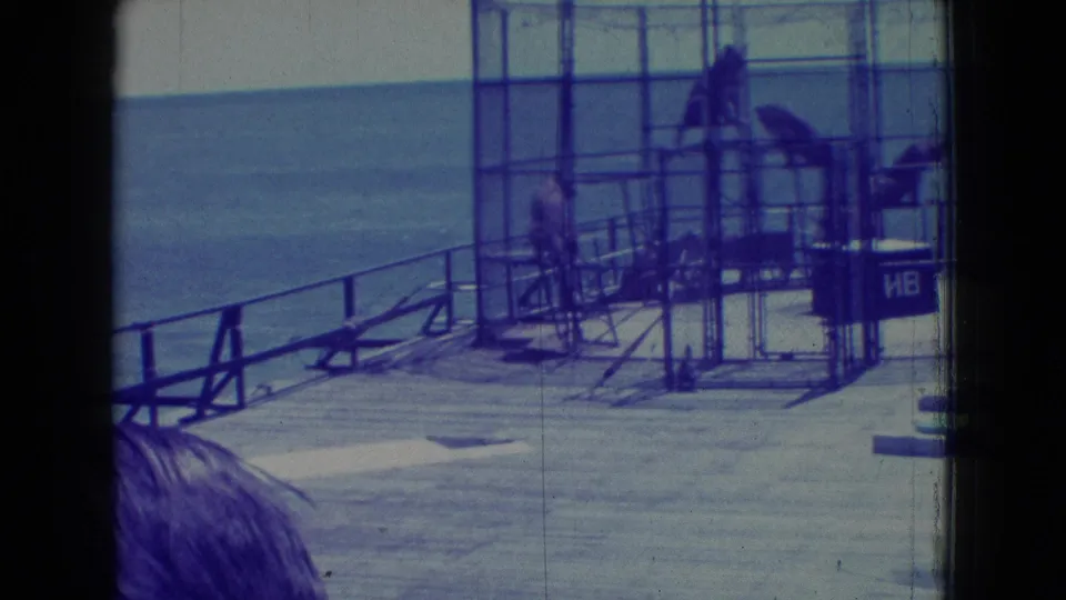 Blue-tinted 1965 New Jersey 8mm frame looking toward a waterfront pier or dock with metal railings, scaffold-like structures, water, and a partially visible person in the foreground.
