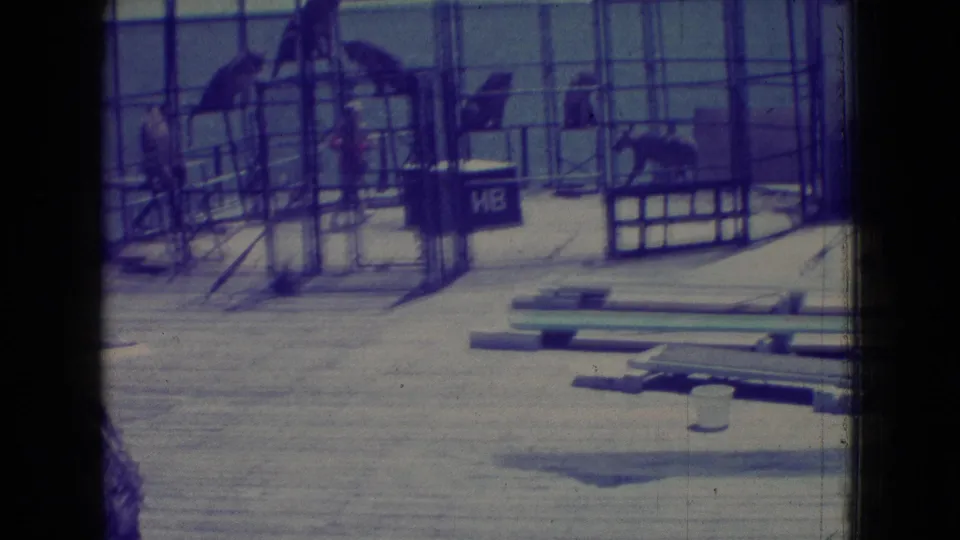 1965 New Jersey home-movie frame showing a seaside pier cage with tigers, a trainer figure, scattered boards in the foreground, and a dark lettered box beside the structure.