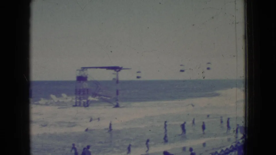 1965 New Jersey 8mm frame showing a wider beachside water view with people wading, distant small boats or objects, and a raised platform structure near the shoreline.