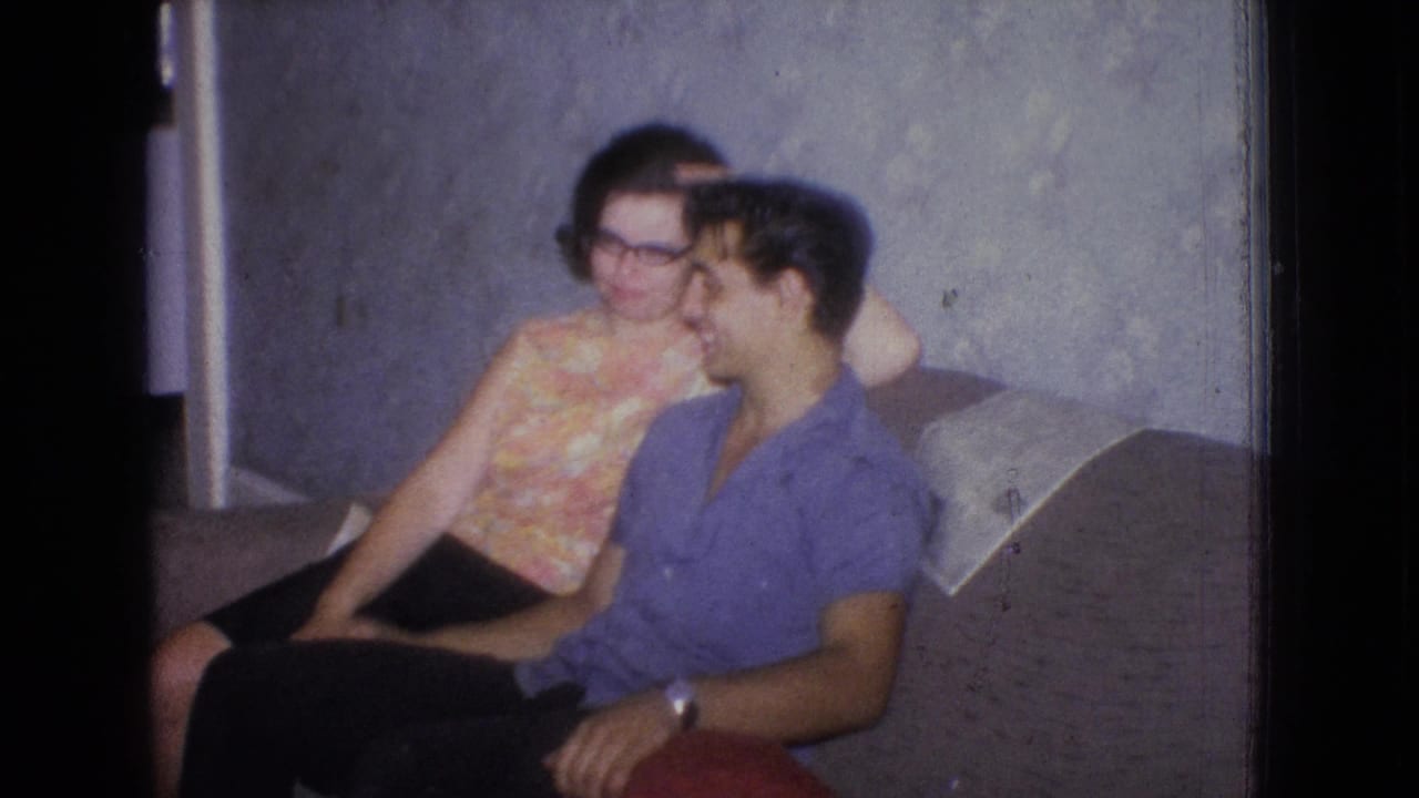 In a 1965 New Jersey home-movie frame, two adults sit close together on a couch against patterned wallpaper in a private living room.