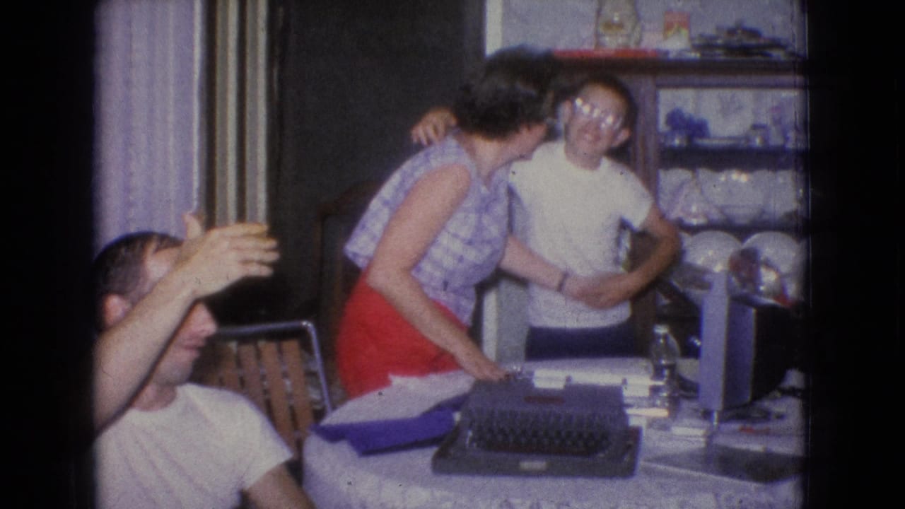 In a 1965 New Jersey domestic interior, a woman in a red skirt embraces a young man near a table with a typewriter as another adult partially blocks the camera.
