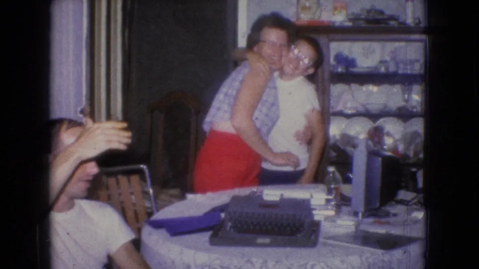 A 1965 New Jersey home-movie frame shows a woman hugging a young man beside a table with a typewriter, with a cabinet of dishes in the background.