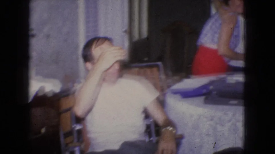 In the closing 1965 New Jersey frame, a seated man covers his face playfully beside a dining table as a woman in a red skirt moves out of view.