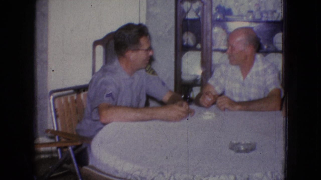 In a 1965 New Jersey home-movie frame, two adult men sit at a covered dining table with a china cabinet and dishes behind them.