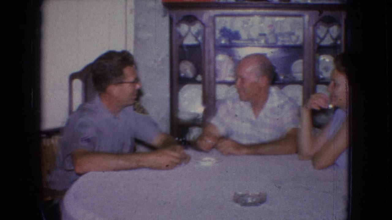 A 1965 New Jersey home-movie frame shows two men and a woman seated around a covered dining table in a private interior with dishes displayed behind them.