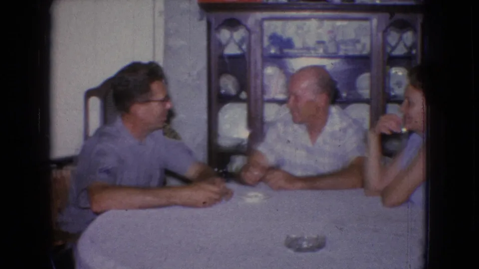 A 1965 New Jersey home-movie frame shows two men and a woman seated around a covered dining table in a private interior with dishes displayed behind them.
