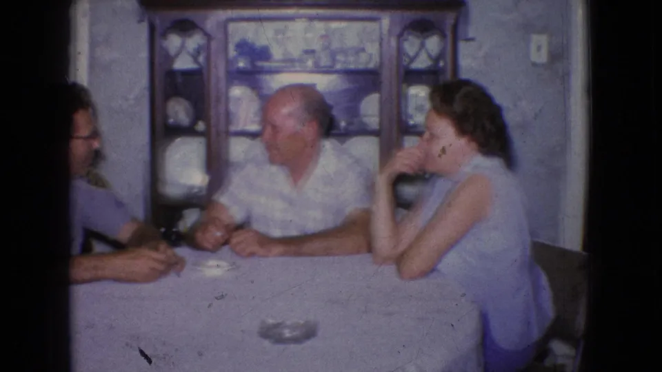 Three adults are seated at a covered dining table in a 1965 New Jersey home interior, with a wooden china cabinet and glass ashtray visible.