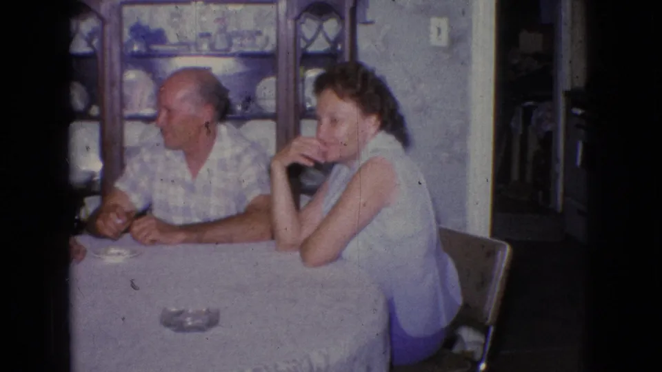 A man in a patterned shirt and a woman in a sleeveless top sit at a covered dining table in a 1965 New Jersey private home interior.
