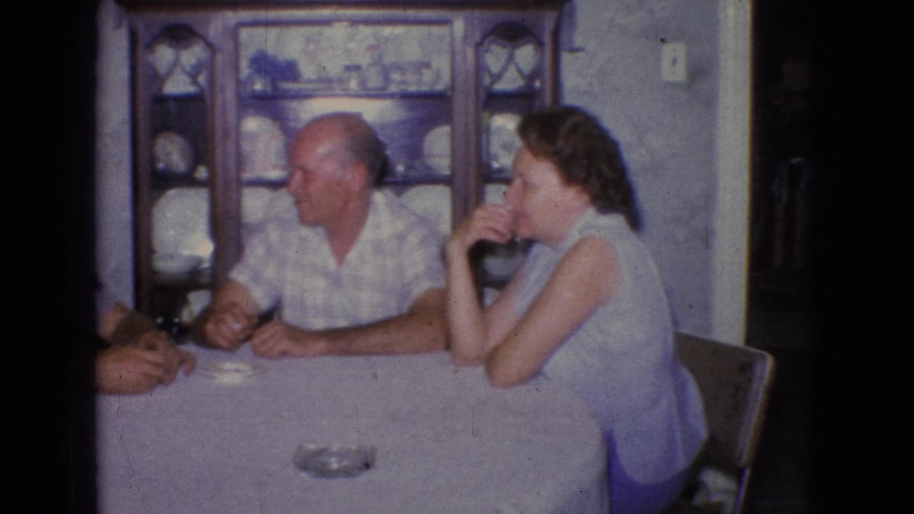 1965 New Jersey 8mm home-movie frame showing three adults seated around a white-covered dining table, with an ashtray and a wooden china cabinet visible in the room.