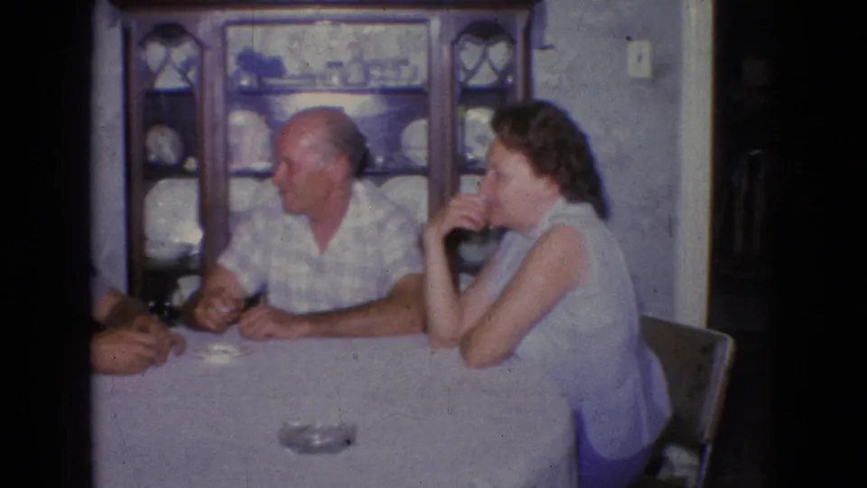 1965 New Jersey 8mm home-movie frame showing three adults seated around a white-covered dining table, with an ashtray and a wooden china cabinet visible in the room.