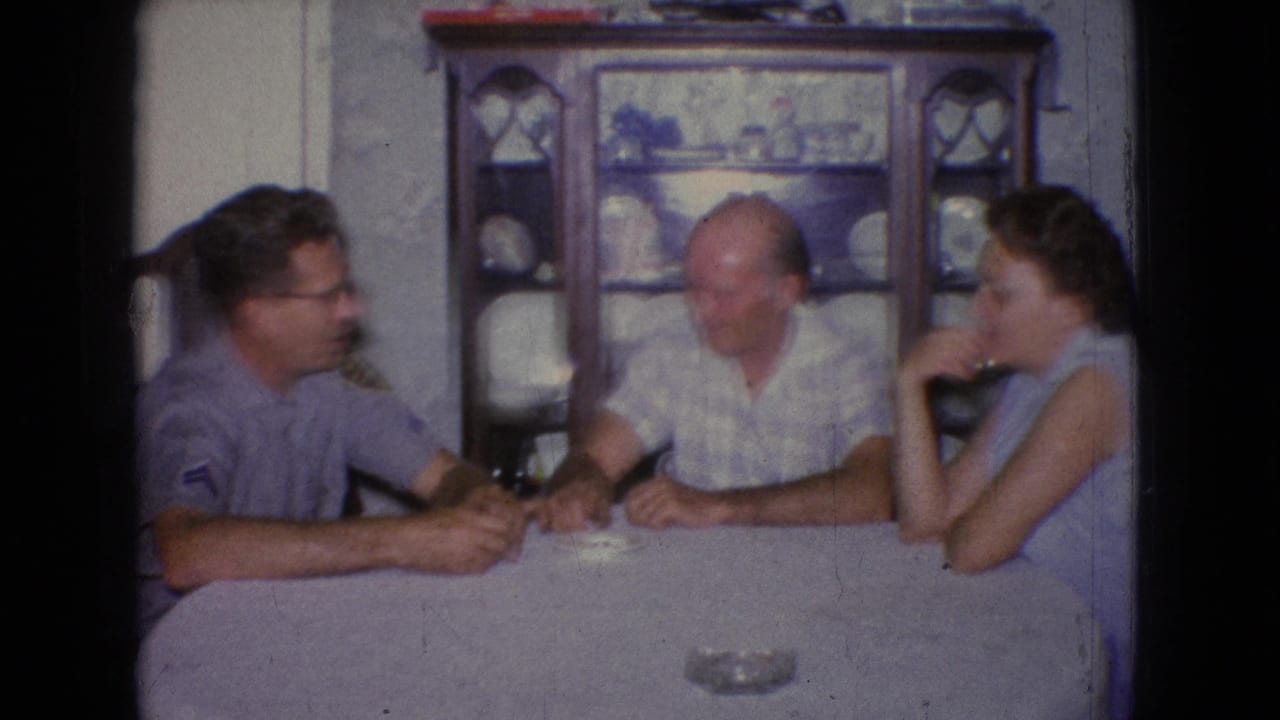 1965 New Jersey home-movie frame with a younger man, an older man, and a woman seated at a dining table in front of a cabinet filled with dishes.