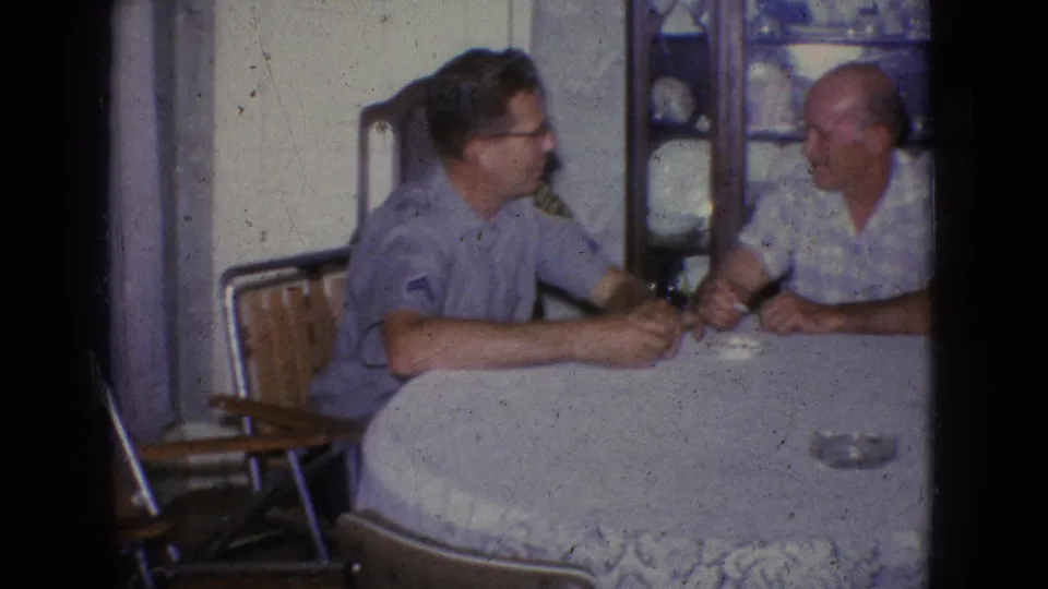 1965 New Jersey home-movie frame showing two men talking at a dining table, with a white tablecloth, ashtray, chairs, and a wooden cabinet visible in the private interior.