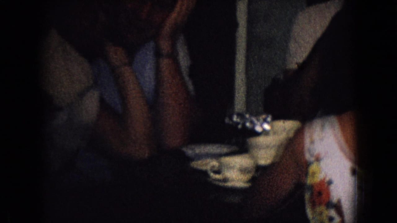 1965 New Jersey home-movie frame showing adults seated close together near a table with teacups and saucers in a dark private interior.