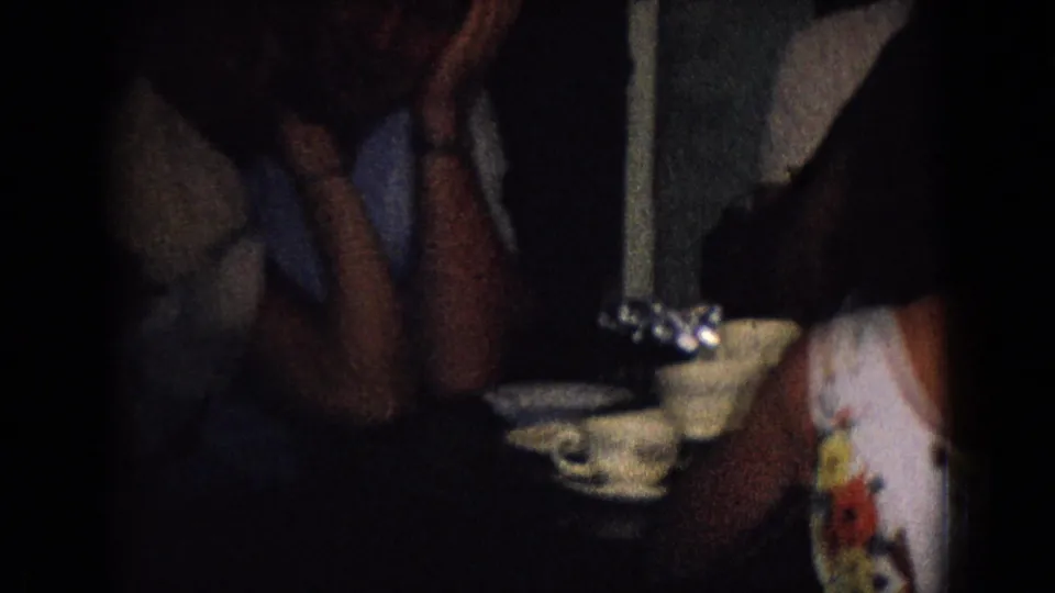 1965 New Jersey home-movie frame showing adults seated close together near a table with teacups and saucers in a dark private interior.