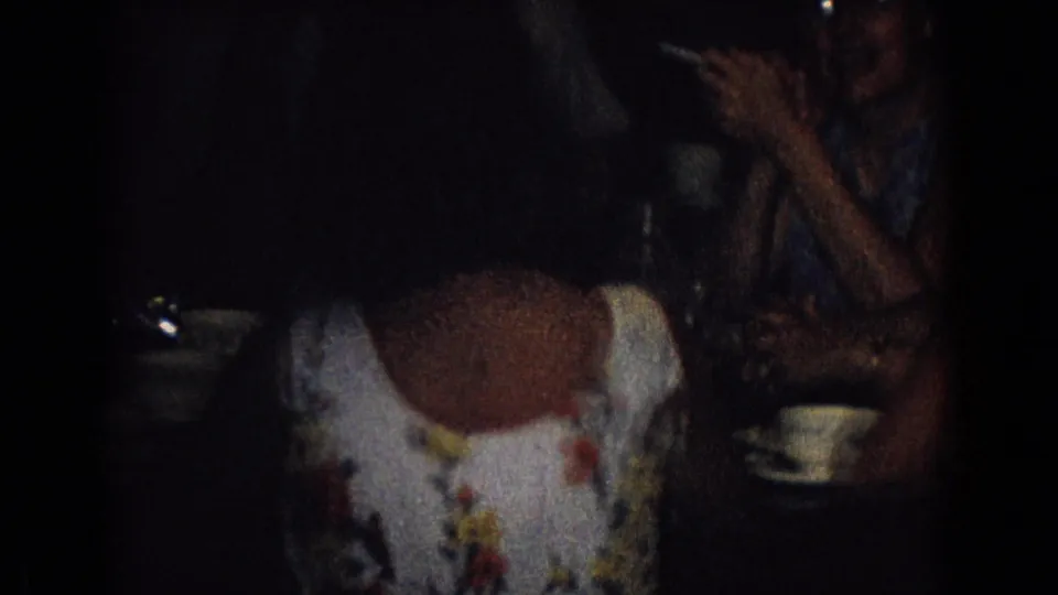 1965 New Jersey home-movie frame showing a woman in a floral dress seated at a table, with a cup and saucer nearby and another adult partly visible in the dark room.