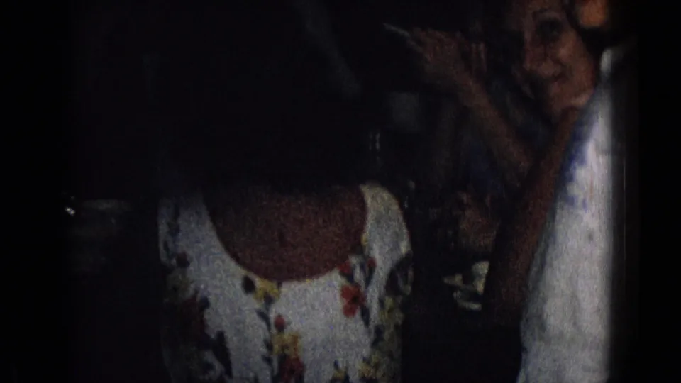 1965 New Jersey home-movie frame showing a woman in a floral dress seated beside another adult at a dimly lit table with cups and saucers visible.