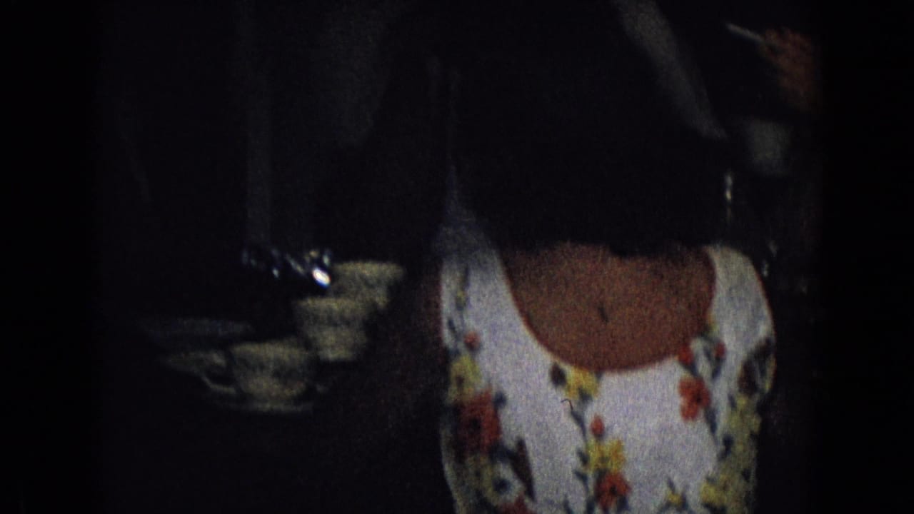 A seated woman in a white floral dress is framed from behind or the side near teacups and saucers in dim 1965 New Jersey home-movie footage.