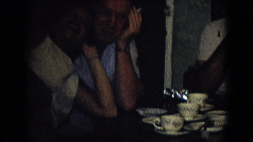 Adults in a dim private interior sit close together around teacups and saucers, with one person raising hands near the face in 1965 New Jersey home-movie footage.
