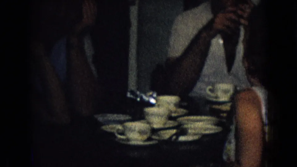 A dim 1965 New Jersey home-movie frame shows seated adults around a table crowded with teacups, saucers, and plates during an informal indoor gathering.