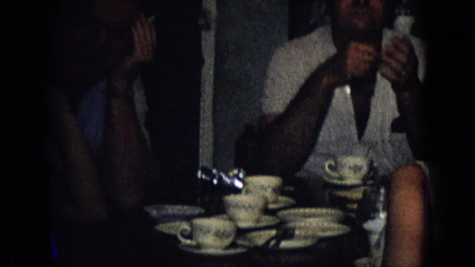 A man in a light shirt sits beside a table of teacups and saucers, with another adult partly visible at the edge of the dim 1965 New Jersey interior frame.
