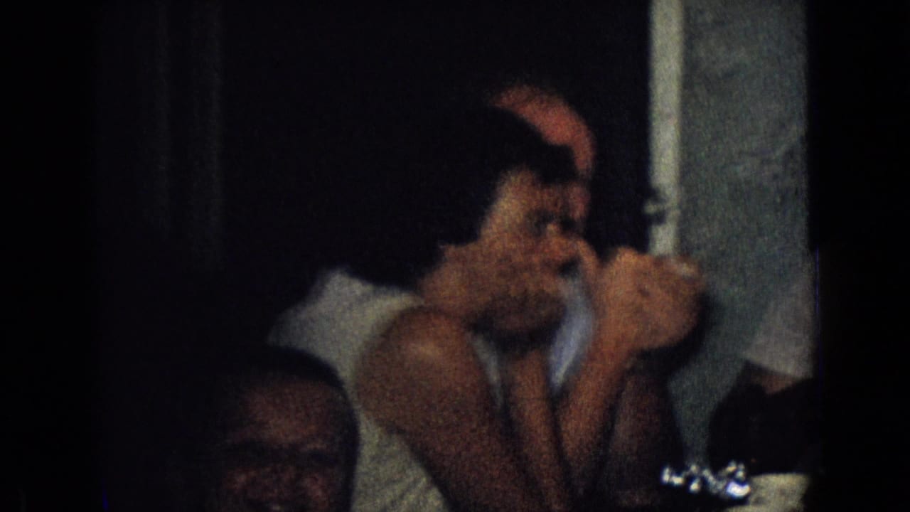 1965 New Jersey home-movie frame showing adults seated close together at a dim indoor dinner table with glassware and dishes in view.