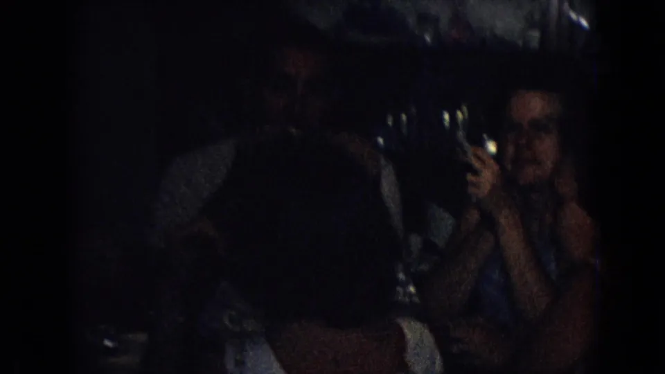 Dim 8mm frame of adults at a dinner table, including a woman holding a cigarette near her face and glassware catching the low light.
