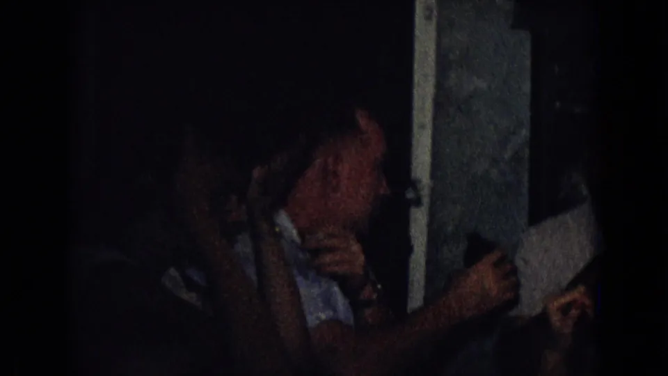 A woman in a sleeveless dress turns toward another guest at a dim indoor dinner table in this 1965 New Jersey home-movie frame.