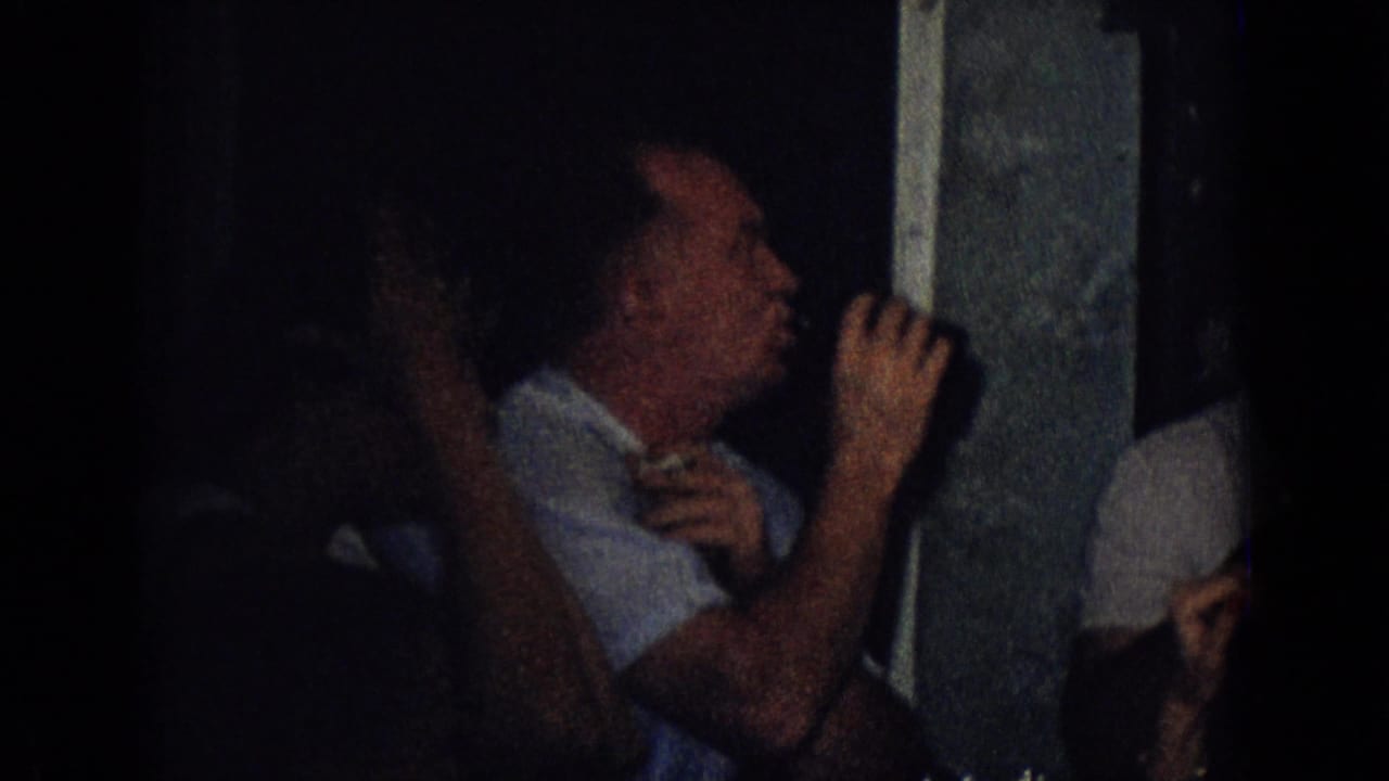 In a dark 1965 New Jersey home-movie party scene, adults sit close together while a man in a light shirt lifts a glass beside a doorway or window frame.