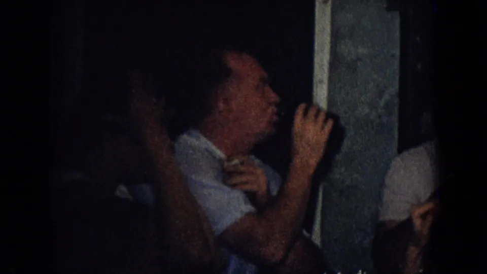 In a dark 1965 New Jersey home-movie party scene, adults sit close together while a man in a light shirt lifts a glass beside a doorway or window frame.