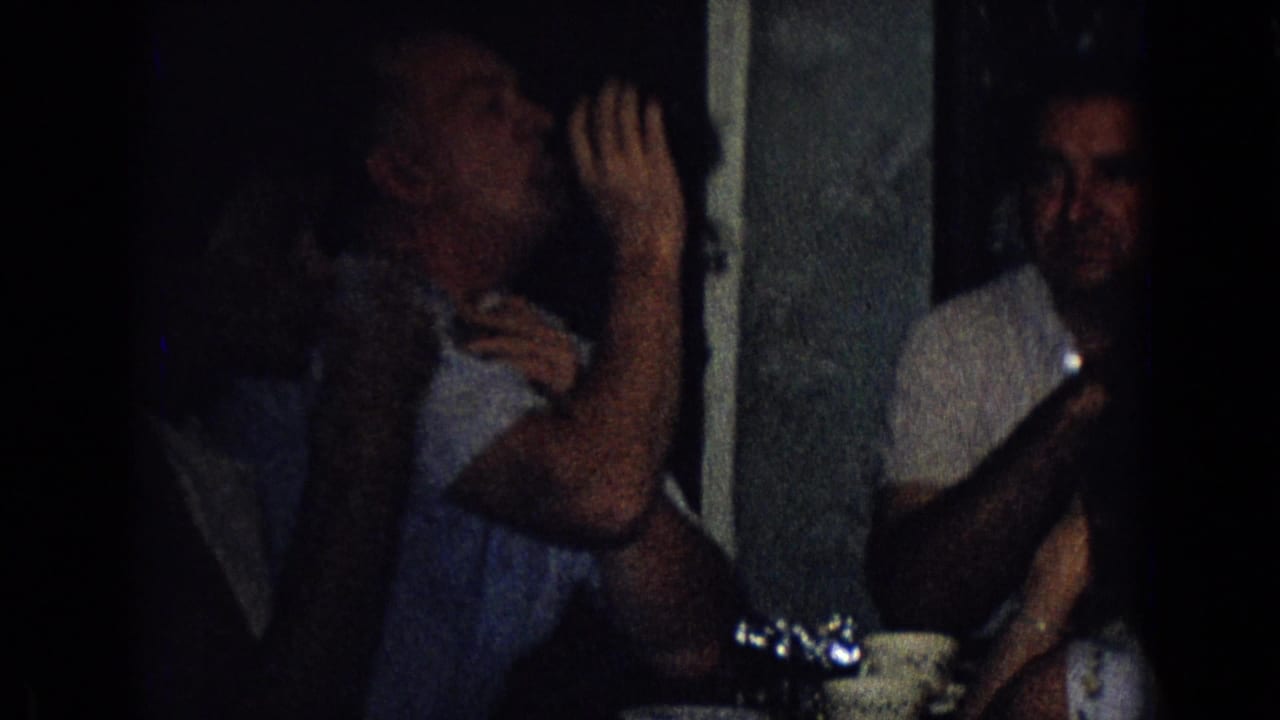 A man drinks from a glass at a dark indoor party in 1965 New Jersey, with seated adults and tableware partly visible around him.